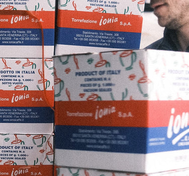Eine Lieferung von Ionia Kartons welche übereinander gestapelt sind, welche gerade ins Geschäft getragen wird. Jeder Karton ist weiß, rot, blau mit dem Ionia Logo in der Mitte. 