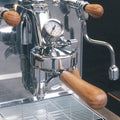 Eine Elba Gentile Espressomaschine aus Edelstahl mit Olivenholzgriffen. In der Mitte ist die Faema E-61 Brühgruppe zu sehen, links davon der Kipphebel für Wasserdampf, rechts, der Kipphebel für Teewasser. Darunter befindet sich die abnehmbare Abtropftasse.