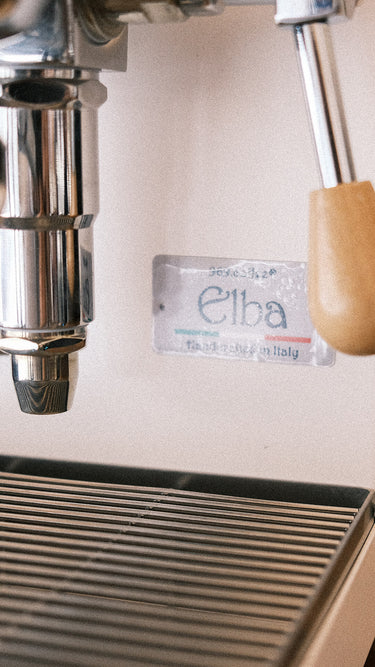 Eine Detailaufnahme der Espressomaschine Elba Mini in weiß mit Olivenholzgriffen. Der Fokus liegt auf dem Schriftzug Elba Handmade in Italy.