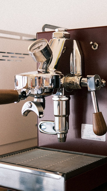Zu sehen ist die Espressomaschine Elba Mini in Bordeaux-Rot mit Sieträgergriff und Kipphebelknauf aus Walnuss Holz. In der Mitte der Brühgruppe befindet sich ein Brühdruck Manometer, auf der linken Seite die einstellbare PID Temperaturkontolle. Darunter die herausziehbare Abtropftasse. Darüber eine Reling um das Herunterfallen der Tassen zu vermeiden.