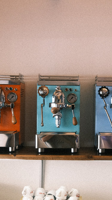 Eine Aufnahme vom Geschäft. Zu sehen sind die Espressomaschinen Elba Quattro in Orange , Türkis und Blau. Darunter ist ein Teil der Tassenablage einer Espressomaschine zu sehen, darauf einige Tassen mit der Aufschrift „Trombetta“ und „Ionia“.