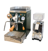 Zu sehen ist die Espressomaschine Elba Quattro in British Racing Green mit Siebträgergriff, Kipphebelknauf und Drehrad für Dampf aus Olivenholz. In der Mitte der Brühgruppe befindet sich ein Brühdruck Manometer, auf der rechten Seite das Kesseldruck-Manometer. Darunter die abnehmbare Abtropftasse. Darüber eine Reling um das Herunterfallen der Tassen zu vermeiden. Daneben ist die On-Demand Kaffeemühle Eureka Mignon Silenzio in chrom abgebildet.