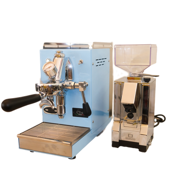 Zu sehen ist die Espressomaschine Elba Mini in hellblau mit Sieträgergriff und Kipphebelknauf aus schwarzem Holz. In der Mitte der Brühgruppe befindet sich ein Brühdruck Manometer, auf der linken Seite die einstellbare PID Temperaturkontolle. Darunter die herausziehbare Abtropftasse. Darüber eine Reling um das Herunterfallen der Tassen zu vermeiden. Daneben ist die On-Demand Kaffeemühle Eureka Mignon Silenzio in chrom abgebildet.