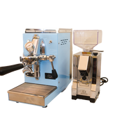 Zu sehen ist die Espressomaschine Elba Mini in hellblau mit Sieträgergriff und Kipphebelknauf aus schwarzem Holz. In der Mitte der Brühgruppe befindet sich ein Brühdruck Manometer, auf der linken Seite die einstellbare PID Temperaturkontolle. Darunter die herausziehbare Abtropftasse. Darüber eine Reling um das Herunterfallen der Tassen zu vermeiden. Daneben ist die On-Demand Kaffeemühle Eureka Mignon Silenzio in chrom abgebildet.