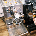 Man sieht die 1-Kreislauf Espressomaschine 969.coffee Elba Mini in Edelstahl. Daneben wird gerade aus der On-Demand Kaffeemühle Eureka Mignon Silenzio Kaffee in einen Siebträger gemahlen. Dahinter sieht man eine Packung Guglielmo Espresso Bar.