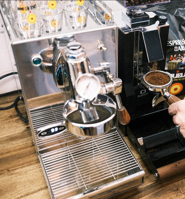 Man sieht die 1-Kreislauf Espressomaschine 969.coffee Elba Mini in Edelstahl. Daneben wird gerade aus der On-Demand Kaffeemühle Eureka Mignon Silenzio Kaffee in einen Siebträger gemahlen. Dahinter sieht man eine Packung Guglielmo Espresso Bar.