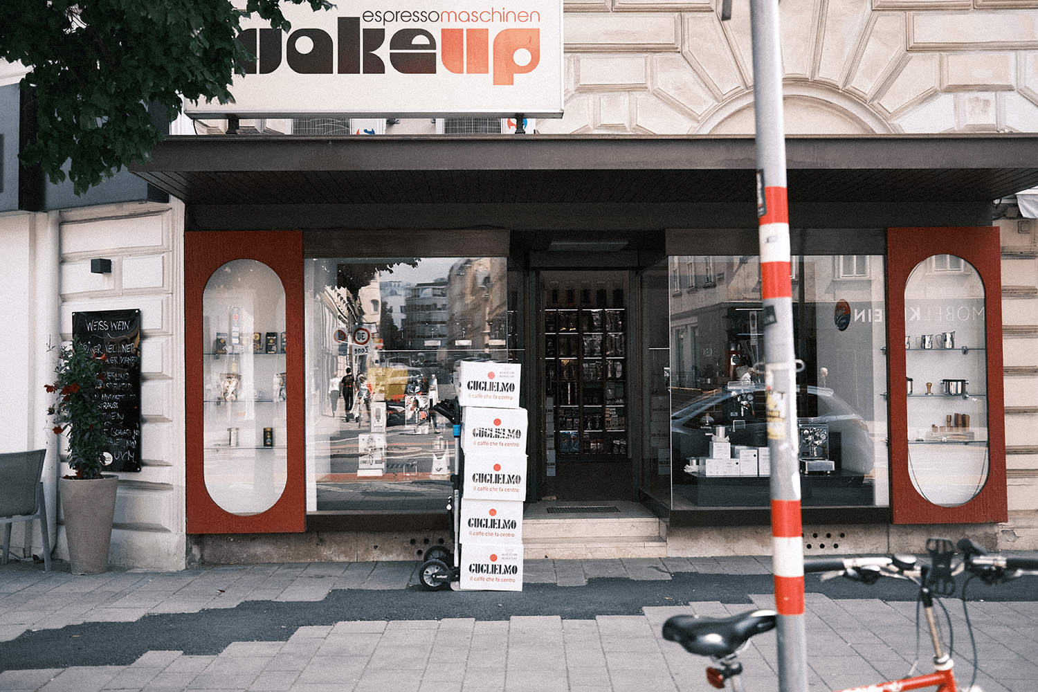 Im Bild ist das Portal von Wake up Espressomaschinen, welches in den Farben rot und braun gehalten ist. Darüber befindet sich ein Vordach. Darüber das große Leuchtschild mit der Logo Wake up Espressomaschinen. Davor ist eine Sackrodel mit mehreren Kisten Kaffee zu sehen.