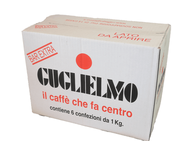 Ein weißer rechteckiger Karton für 6x1kg Bohnenkaffee Guglielmo Extra Bar. Auf der Vorderseite in der Mitte befindet sich der schwarze Guglielmo Schriftzug. Über dem I ist ein großer roter Punkt abgebildet, darunter il caffè che fa centro.  Darunter die Angabe: contiene 6 confezioni da 1kg. Rechts oben in roter, leicht schräger Schrift Bar Extra.