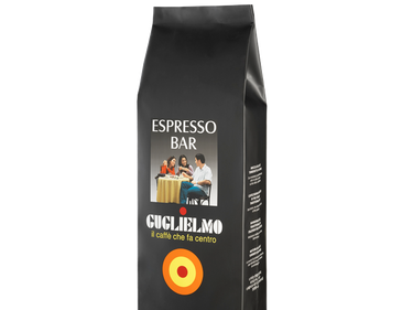 Eine schwarze Aromaverpackung für den Kaffee Guglielmo Espresso Barr in Bohnen.  Oben ist der weiße Schriftzug Espresso Bar zu lesen. In der Mitte ein Bild von 2 Frauen und einem Mann die an einem Tisch sitzen und Kaffee trinken. Darunter der Schriftzug Guglielmo il caffè che fa centro. Unten sieht man das Logo mit dem orange, gelb, roten konzentrischen Kreis.