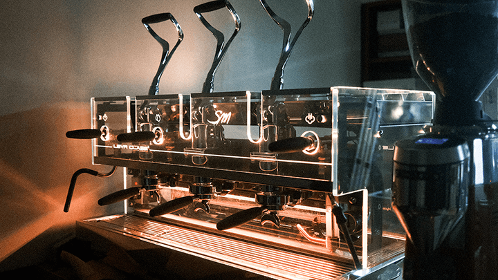 Eine Gesamtansicht der 3-gruppigen Handhebel Kaffeemaschine SM Leva Luxury. Die Maschine ist komplett aus Glas verkleidet und in diesem Bild gelb beleuchtet.