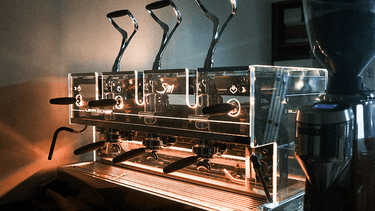 Eine Gesamtansicht der 3-gruppigen Handhebel Kaffeemaschine SM Leva Luxury. Die Maschine ist komplett aus Glas verkleidet und in diesem Bild gelb beleuchtet.