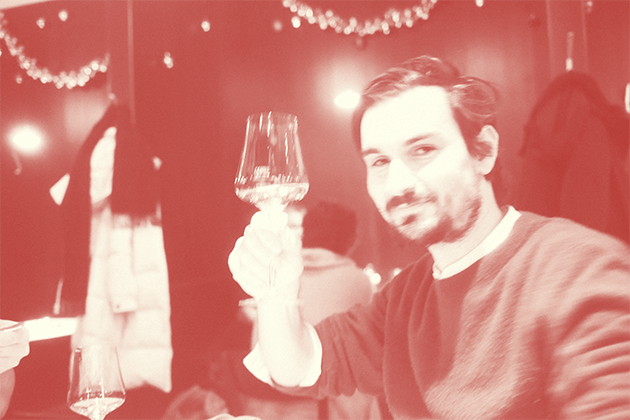 Zu sehen ist ein Foto mit Rotfilter von Kenzo Agostini. Es wurde bei einer Weihnachtsfeier aufgenommen. Kenzo trägt einen dunklen Pullover, in seiner rechten Hand hält er ein Glas Weißwein. 