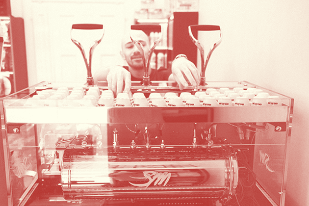 Zu sehen ist ein Foto mit Rotfilter von Mario Sciurti. Im Vordergrund ist die Espressomaschine La San Marco Leva Luxury  mit Ihrer beleuchteten Glasverkleidung zu sehen. Darüber die drei Handhebel. Mario steht dahinter und nimmt gerade 2 Tassen von der Tassenablage um einen Kaffee zuzubereiten. 