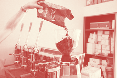 Zu sehen ist eine Aufnahme mit Rotfilter. Dabei wird gerade eine Packung Guglielmo Caffetteria Kaffeebohnen in die Mazzer Robur S Kaffeemühle geleert. Links daneben ist die 3-gruppige Handhebel Espressomaschine Izzo Valchiria zu erkennen.