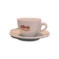 Eine weiße dickwandige Cappuccinotasse mit Unterteller mit dem Henkel auf der rechten Seite. In der Mitte der Tasse ist der rote Trombetta Schriftzug abgebildet.
