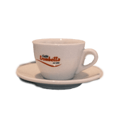 Eine weiße dickwandige Cappuccinotasse mit Unterteller mit dem Henkel auf der rechten Seite. In der Mitte der Tasse ist der rote Trombetta Schriftzug abgebildet.