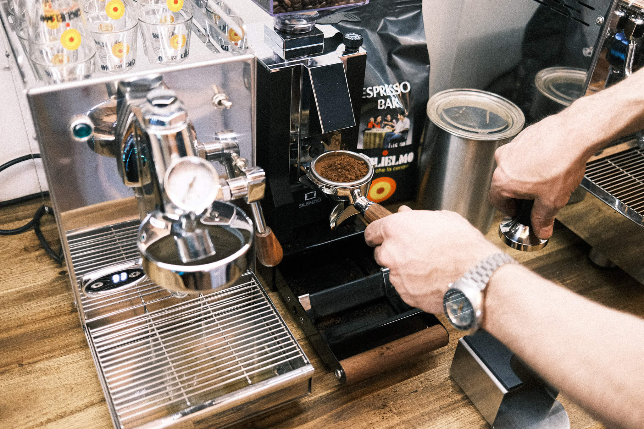 Man sieht die 1-Kreislauf Espressomaschine 969.coffee Elba Mini in Edelstahl. Daneben wird gerade aus der On-Demand Kaffeemühle Eureka Mignon Silenzio Kaffee in einen Siebträger gemahlen. Dahinter sieht man eine Packung Guglielmo Espresso Bar neben einem Vakuumbehälter zur Aufbewahrung der Bohnen.