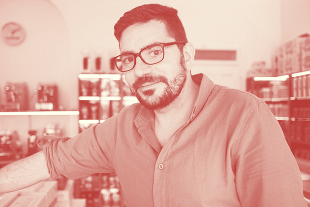 Zu sehen ist ein Foto mit Rotfilter von Viktor Alonso Fox. Es wurde im Geschäft aufgenommen. Er trägt ein Hemd mit aufgekrempelten Ärmeln, im Hintergrund ist das Regal mit einer Auswahl von Bohnenkaffees zu sehen.
