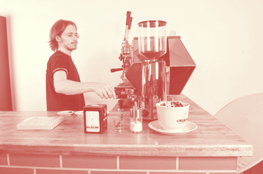 In dieser Aufnahme mit Rot-Filter aus dem Jahr 2016 sieht man den Chef bei der Bedienung unserer früheren Espressomaschine La Pavoni Diamante mit 2 Hebelbrühgruppen. Auf der Bar ist eine große Tasse mit Zuckerportionen darin zu sehen, daneben ein Zuckerstreuer, ein Glas mit Löffeln sowie ein Serviettenhalter mit Guglielmo Logo. Es wird gerade mit der Eureka Zenith 65E Kaffeemühle eine doppelte Espressoportion gemahlen.