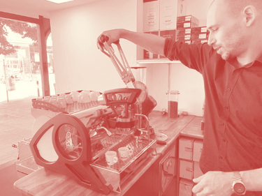 In dieser Aufnahme mit Rot-Filter aus dem Jahr 2021 sieht man den Chef bei der Bedienung unserer früheren Espressomaschine La Marzocco Leva mit 1 Hebelbrühgruppe. Die Maschine ist in Richtung der Auslage aufgestellt, darauf sind einige Guglielmo Espresso- und Cappuccinotassen zu sehen. Im Bild wird gerade der Hebel hinuntergezogen um einen Leerbezug zu machen.